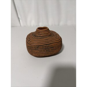 May include: A small, squat, terracotta-colored ceramic vase with a rounded, flattened shape. The surface features a series of horizontal, incised dark lines that wrap around the body and the narrow, raised neck, creating a textured, layered appearance.