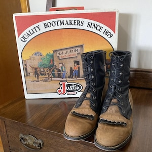 Könnte beinhalten: Ein Paar Schnürstiefel mit beigefarbenen Wildledervorderkappen und schwarzen Lederschäften mit schwarzen Fransen. Die Stiefel stehen vor einem weißen Karton mit der Aufschrift QUALITY BOOTMAKERS SINCE 1879 und einer Laden-Illustration.