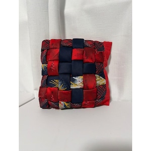 May include: A square fabric coaster or small mat featuring a woven design of navy blue strips and red strips with printed patterns of small white Christmas trees, gold stars, and plaid textures. The edges are finished with a red fabric border.