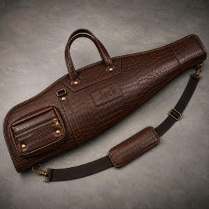 Pode incluir: Um estojo de transporte castanho escuro com relevo de crocodilo, com uma pega superior e uma alça de ombro preta removível. O saco apresenta um bolso frontal retangular com fecho, ferragens em tom de latão e o nome Jack gravado.