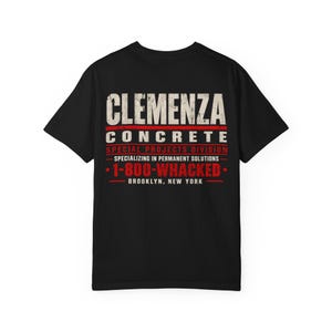 Puede incluir: Una camiseta negra de manga corta con texto desgastado en blanco y rojo en la parte posterior que dice CLEMENZA CONCRETE SPECIAL PROJECTS DIVISION SPECIALIZING IN PERMANENT SOLUTIONS 1-800-WHACKED BROOKLYN, NEW YORK con líneas rojas.
