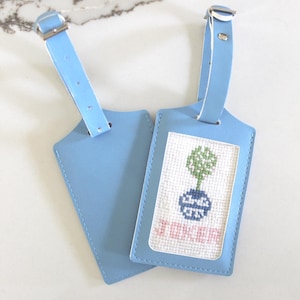 May include: Two light blue luggage tags with adjustable straps and silver buckles. One tag features a white rectangular insert with a cross-stitched design of a green plant in a blue pot above the pink embroidered text JOKER.