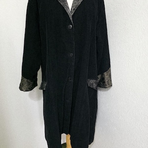 May include: A long black coat featuring a dark grey floral patterned collar and matching cuffs. The coat has a button-down front closure with four visible buttons and long sleeves. The fabric appears to have a textured finish throughout the main body.