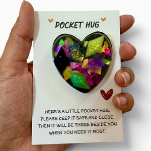 May include: A heart-shaped resin charm filled with colourful glitter and geometric shards, attached to a white card printed with the text POCKET HUG and a poem that reads, Here's a little pocket hug, please keep it safe and close.