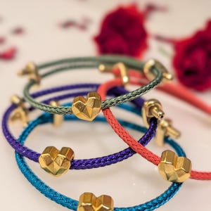 Peut inclure: Un ensemble de quatre bracelets en cordon tressé de couleur vert olive, rouge vif, violet profond et bleu cyan. Chaque bracelet comporte une breloque en forme de cœur facetté doré et un petit fermoir métallique de couleur dorée.