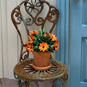 Può includere: Una sedia in metallo arrugginito con un designo ornato si trova su un patio in pietra. Un vaso in terracotta con fiori arancioni è posto sul sedile della sedia. La sedia è posizionata davanti a una porta blu con una maniglia dorata.