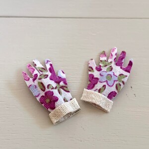 Può includere: Un paio di piccoli guanti da giardinaggio con un motivo floreale nei toni del viola, verde e blu su sfondo bianco. I polsini sono in tessuto beige strutturato. I guanti sono appoggiati su una superficie di legno chiaro.