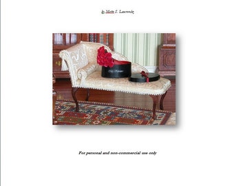 Op de afbeelding: Een miniatuur witte chaise longue met een boeket rode rozen in een zwarte geschenkdoos met de tekst 'My Valentine'. De chaise longue staat op een rood, blauw en wit gepatroneerd tapijt.