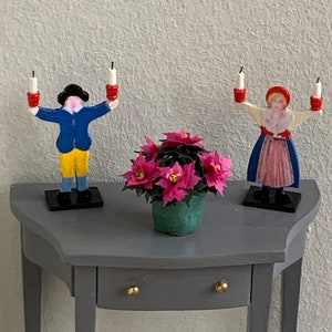 Könnte beinhalten: Ein grauer Tisch mit einer Schublade und zwei Kerzenständern aus Holz. Die Kerzenständer sind bemalt, um wie Menschen auszusehen, die Kerzen halten. Eine Person trägt eine blaue Jacke und eine gelbe Hose, die andere trägt ein rotes Kleid. Eine Topfpflanze mit rosa Blüten steht auf dem Tisch.