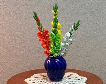 May include: A blue ceramic vase with a swirl design holds a bouquet of paper flowers. The flowers are orange, yellow, white, and green. The vase is sitting on a wooden table with a white crocheted doily.
