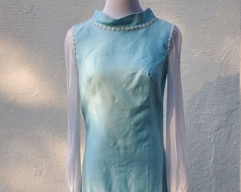 May include: A light blue maxi dress with long sheer sleeves and a beaded neckline. The dress is on a white mannequin against a white wall.