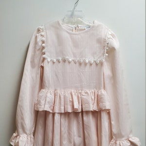 Puede incluir: Un vestido de rayas rosa y blancas con un dobladillo con volantes y mangas largas. El vestido tiene un ribete de encaje blanco alrededor del escote y un botón blanco en la espalda.