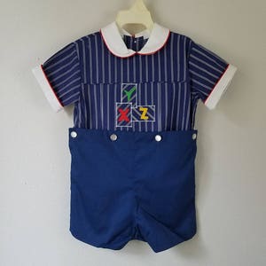 May include: A blue and white striped short-sleeved shirt with a white collar and red trim. The shirt has a blue, red, yellow, and green embroidered design of blocks with the letters X, Y, and Z on the front. The shirt is paired with blue shorts.