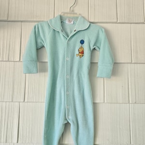 May include: A light blue baby onesie with long sleeves and attached feet, hanging on a hanger. The onesie features a collar and a button-up front. An embroidered image of Winnie the Pooh holding a blue balloon is on the chest.