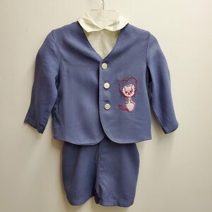 Puede incluir: Un traje azul vintage para niño, compuesto por una chaqueta y un pantalón corto. La chaqueta tiene un cuello de lunares blancos y un diseño de león bordado en rojo y blanco. El traje está abotonado y colgado de una percha.