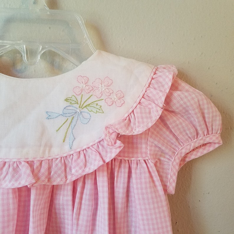 K&ouml;nnte beinhalten: Ein rosa-wei&szlig;es Vichy-Kleid mit einem wei&szlig;en Kragen mit gesticktem Blumendesign. Das Kleid hat kurze, gepuffte &Auml;rmel und einen R&uuml;schenkragen. Das Kleid h&auml;ngt an einem durchsichtigen Kunststoffb&uuml;gel.