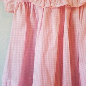 K&ouml;nnte beinhalten: Nahaufnahme eines rosa-wei&szlig; karierten Kleides. Das Kleid hat einen ger&uuml;schten Ausschnitt und kurze &Auml;rmel. Der Stoff ist weich und flie&szlig;end, mit vertikalen Falten, die Schatten erzeugen. Das Kleid ist wahrscheinlich f&uuml;r ein Kind.