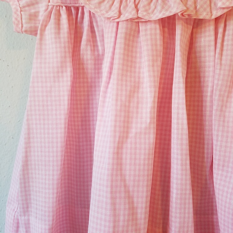 K&ouml;nnte beinhalten: Nahaufnahme eines rosa-wei&szlig; karierten Kleides. Das Kleid hat einen ger&uuml;schten Ausschnitt und kurze &Auml;rmel. Der Stoff ist weich und flie&szlig;end, mit vertikalen Falten, die Schatten erzeugen. Das Kleid ist wahrscheinlich f&uuml;r ein Kind.