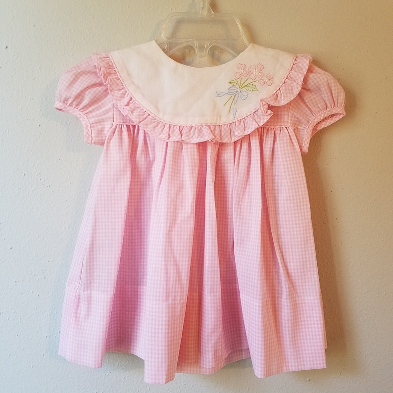 K&ouml;nnte beinhalten: Ein rosa-wei&szlig;es Vichy-Kleid mit wei&szlig;em Kragen und kurzen &Auml;rmeln. Der Kragen ist mit R&uuml;schen verziert und hat ein gesticktes Blumendesign mit einer blauen Schleife. Das Kleid h&auml;ngt an einem durchsichtigen Kunststoffb&uuml;gel.