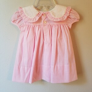K&ouml;nnte beinhalten: Ein rosa-wei&szlig; kariertes Babykleid mit wei&szlig;em Bubikragen und kurzen, gepufften &Auml;rmeln. Das Kleid hat einen Knopfverschluss am R&uuml;cken und h&auml;ngt an einem durchsichtigen Kunststoffb&uuml;gel.