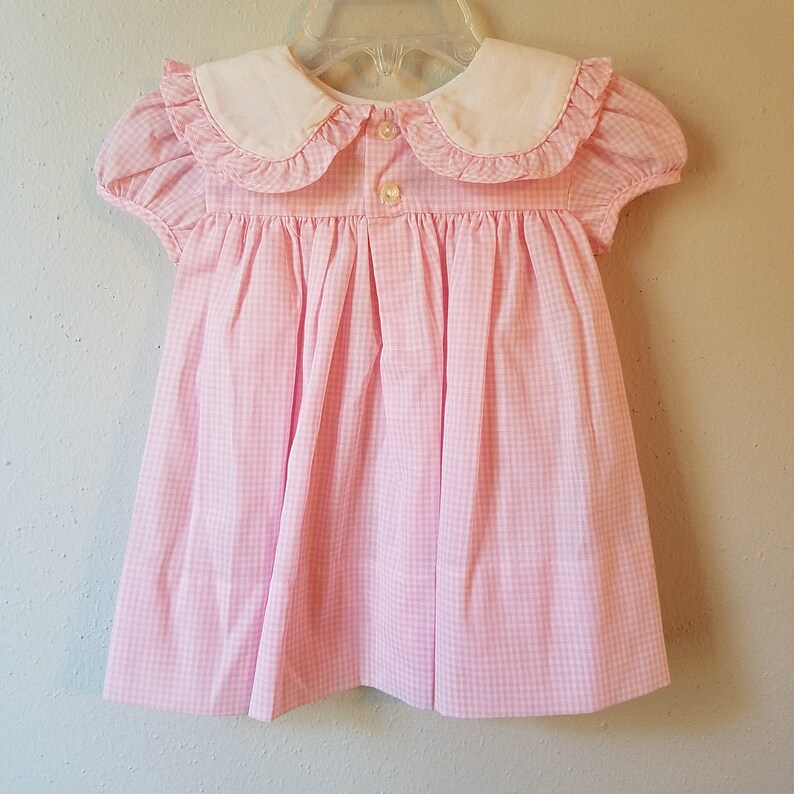 K&ouml;nnte beinhalten: Ein rosa-wei&szlig; kariertes Babykleid mit wei&szlig;em Bubikragen und kurzen, gepufften &Auml;rmeln. Das Kleid hat einen Knopfverschluss am R&uuml;cken und h&auml;ngt an einem durchsichtigen Kunststoffb&uuml;gel.