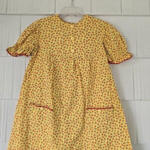 May include: Yellow floral print dress with short sleeves and red trim. The dress has a round neckline, button details, and two front pockets with red trim. The dress is hanging on a white hanger.