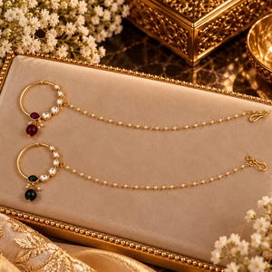 May include: Two gold-toned nose rings featuring circular hoops adorned with clear stones. One ring has a deep red teardrop stone and a round red bead, while the other has a green teardrop stone and a black bead. Both include a delicate pearl chain.