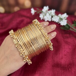 May include: A set of gold-coloured bangles featuring a stack of thin, smooth circular bands. The two outer bangles are designed with a decorative beaded texture, while the inner bangles remain plain and polished, creating a uniform metallic appearance.