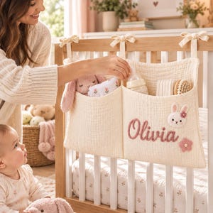 May include: A cream-colored fabric crib organizer with two pockets, featuring the name Olivia embroidered in dusty pink script, a small crocheted bunny face, and a pink flower appliqué. The organizer hangs from the crib rail with fabric ties.
