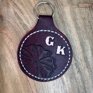 May include: A circular dark purple leather keychain featuring a stamped floral design on the left side and the embossed letters G and K on the right. The edges are secured with thick white stitching, and a silver metal ring is attached at the top.