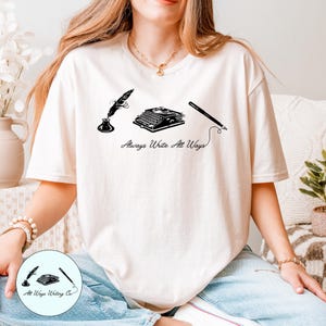 May include: A cream-colored short-sleeve t-shirt featuring a black graphic print of a quill in an inkwell, a vintage typewriter, and a fountain pen. Below the illustrations, the text reads Always Write All Ways in a cursive script font.