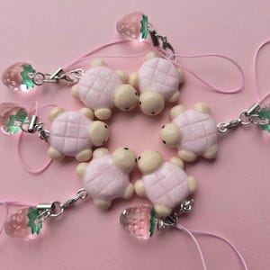 May include: A collection of five charms featuring light pink turtle figures with glittery shells and cream-colored heads. Each turtle is attached to a pink cord and a small, translucent pink strawberry charm with a green leaf top via a silver clasp.
