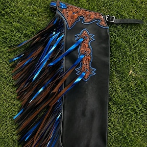 Pode incluir: Um chap de couro preto com detalhes em couro castanho com gravação floral e costuras em azul brilhante. O lado é adornado com longas tiras de franjas em camadas alternando as cores preto, castanho e azul metálico, preso por uma fivela preta.