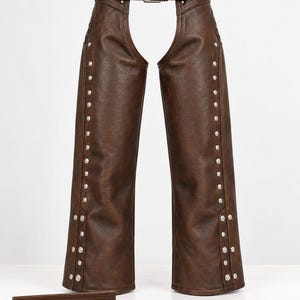 May include: Dark brown leather chaps featuring a waist belt with a metal buckle, silver-toned metal studs running down the outer legs, and a small rectangular wooden sign at the base with the text CRAFTED LEGACY printed in gold lettering.