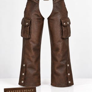 May include: Dark brown leather chaps featuring a waist belt with a metal buckle, two large cargo pockets with snap closures on the thighs, and three metal snaps along the lower outer leg. A small rectangular sign reads CRAFTED LEGACY.