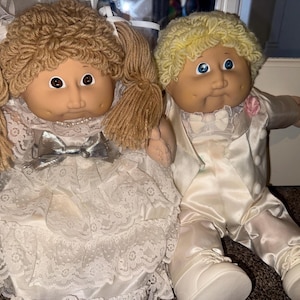 May include: Two dolls with yarn hair and dimpled faces. The left doll has brown yarn hair in pigtails and wears a white lace dress with a silver bow. The right doll has blonde yarn hair and wears a white satin suit with a small pink flower.