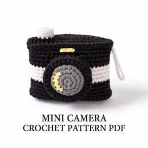 Può includere: Una fotocamera giocattolo all'uncinetto nera, bianca, grigia e gialla con obiettivo centrale, pulsante superiore e cinturino laterale. Il testo sottostante recita MINI CAMERA CROCHET PATTERN PDF. L'oggetto presenta punti di lana visibili.