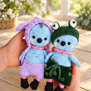 Può includere: Due bambole all'uncinetto fatte a mano con corpo azzurro, occhi neri e guance rosa. Una indossa un completo color lavanda con cappuccio e stivali neri, l'altra un cappello a forma di rana verde scuro e pantaloncini abbinati con stivali.
