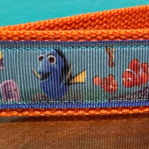 May include: Orange fabric keychain with a blue ribbon featuring characters from the movie "Finding Nemo." The keychain has a silver metal ring and a gold-colored metal clasp.