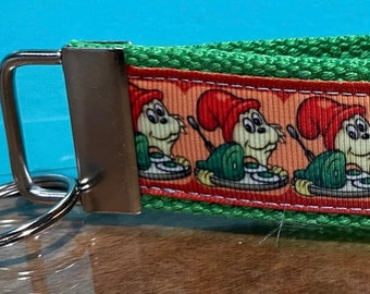 Puede incluir: Un llavero de pulsera verde y naranja. La pulsera está hecha de tela verde tejida con una cinta estampada con personajes de dibujos animados con sombreros rojos y tocadiscos verdes. Se adjunta un cierre y un llavero de metal plateado.