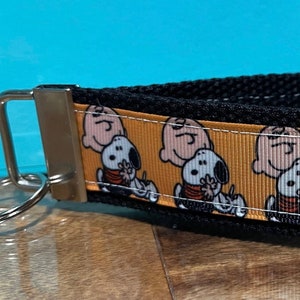 May include: A black wrist lanyard with a metal clasp and key ring. The lanyard features a repeating pattern of Charlie Brown and Snoopy on an orange background. The lanyard is made of a durable woven material.