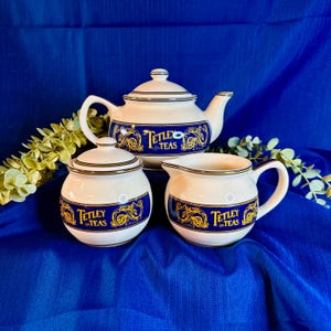 May include: A ceramic tea set consisting of a teapot, a sugar bowl with a lid, and a creamer. Each piece is cream-coloured with a wide blue band featuring gold scrollwork and the text Tetley Teas. The set has thin gold trim on the rims and lids.
