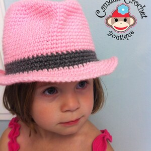 May include: A young girl wearing a pink and gray crocheted hat. The hat has a wide brim and a gray band around the crown. The hat is made with a single crochet stitch.
