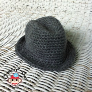 May include: A gray crocheted fedora hat with a wide brim. The hat is sitting on a white wicker surface.