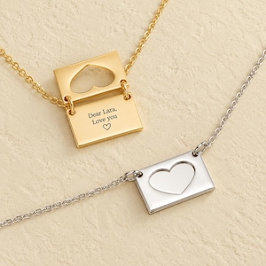 Könnte beinhalten: Eine goldfarbene Halskette mit einem rechteckigen Umschlaganhänger, der einen Herzausschnitt und eine offene Klappe mit der Gravur Dear Lara, Love you sowie einem Herzsymbol aufweist, neben einer silberfarbenen Kette mit gleichem Anhänger.