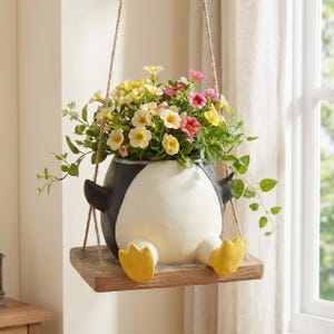 Puede incluir: Una maceta colgante con forma de pingüino, cuerpo blanco y negro y pies amarillos, sobre una plataforma de madera rectangular suspendida por cuerdas de yute. La maceta contiene una variedad de pequeñas flores amarillas, rosas y blancas.