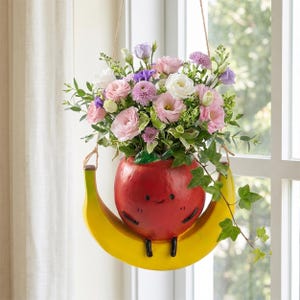 Puede incluir: Una maceta colgante con forma de tomate rojo sentado sobre un plátano amarillo en forma de media luna. El tomate tiene un rostro negro sencillo y está lleno de un ramo de flores rosas, blancas y moradas con hojas de hiedra verde.