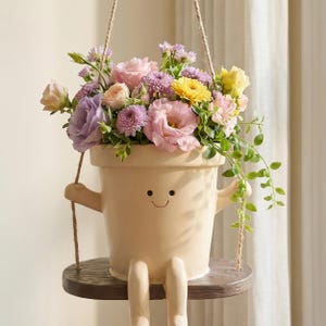 Puede incluir: Una maceta de cerámica beige con forma de persona con cara sonriente, sentada en un pequeño columpio de madera marrón. La maceta está llena de un colorido ramo de flores rosas, moradas y amarillas, incluyendo rosas y crisantemos con hojas verdes.
