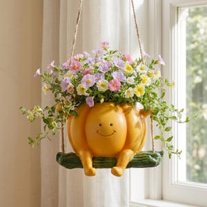 Puede incluir: Una maceta con forma de calabaza amarilla con una cara sonriente, sentada sobre un columpio con forma de rama verde suspendido por cuerdas de yute. La maceta está llena de un denso arreglo de pequeñas flores de petunia multicolores.