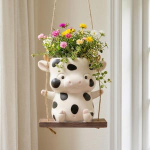 Puede incluir: Una maceta colgante de cerámica con forma de vaca con manchas negras sobre un cuerpo color crema. La maceta descansa sobre un pequeño columpio de madera suspendido por cuerdas de yute y está llena de pequeñas flores rosas, amarillas y blancas.